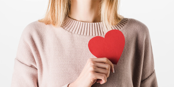 Eine Person hält ein rotes, ausgeschnittenes Papierherz vor der Brust. Der Oberkörper ist in einem hellen Strickpullover zu sehen, der Hintergrund ist hell.