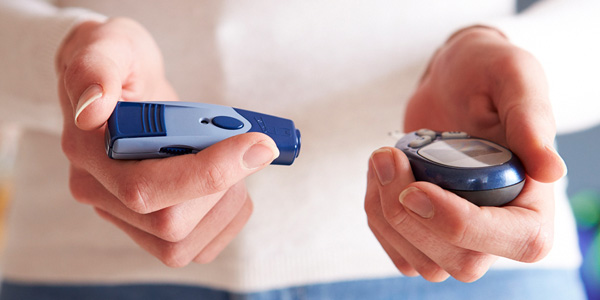 Zwei Hände halten ein kleines rundes Messgerät und ein längliches blaues Stechgerät zur Blutzuckermessung.
